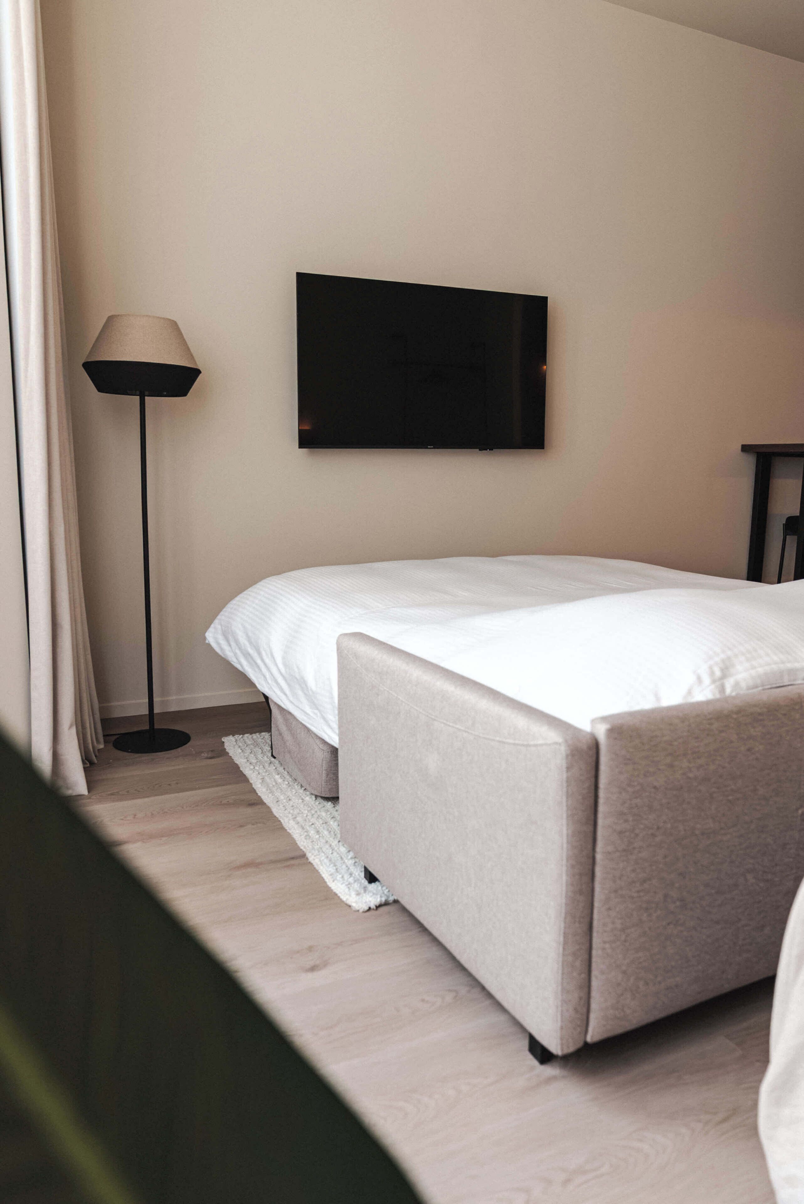 A modern hotel room with a bed, a flat-screen TV mounted on the wall, a floor lamp, and a small table.