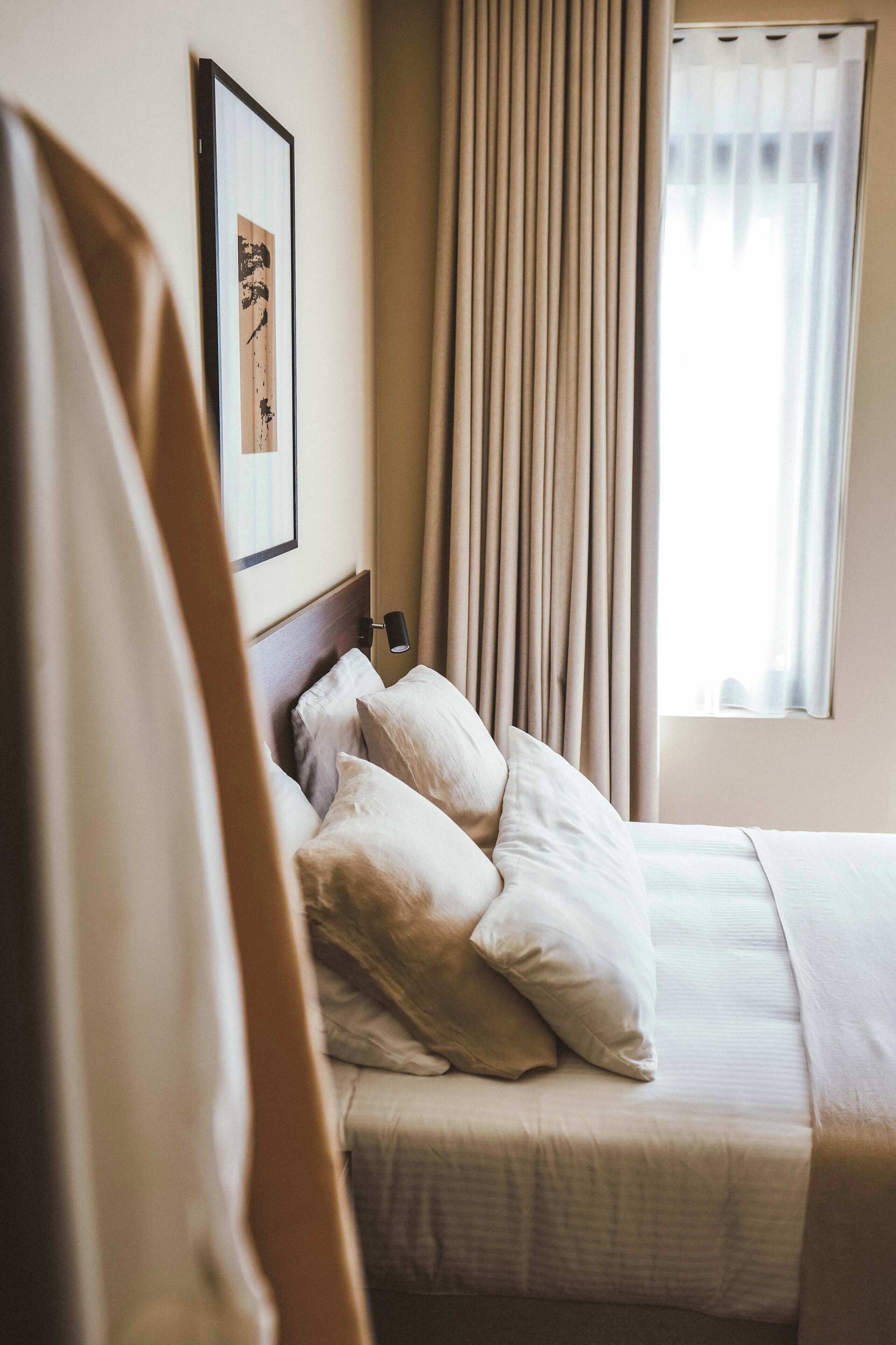 Une chambre d’hôtel avec un lit, des oreillers, une tête de lit, une table de chevet, une lampe, une fenêtre avec des rideaux et un tableau encadré au mur.