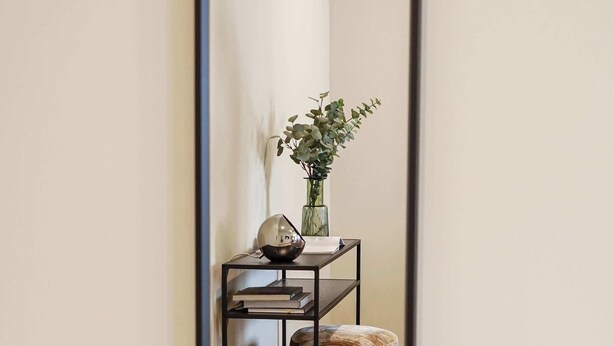 Un espejo refleja una habitación con suelo de madera, un estante metálico negro con libros y un jarrón, y un taburete pequeño.