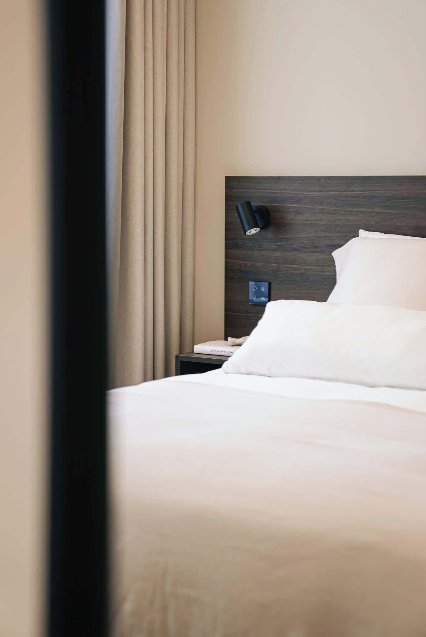 Habitació d'hotel amb llit, una tauleta de nit, una làmpada i un panell de control fixat a la paret.
