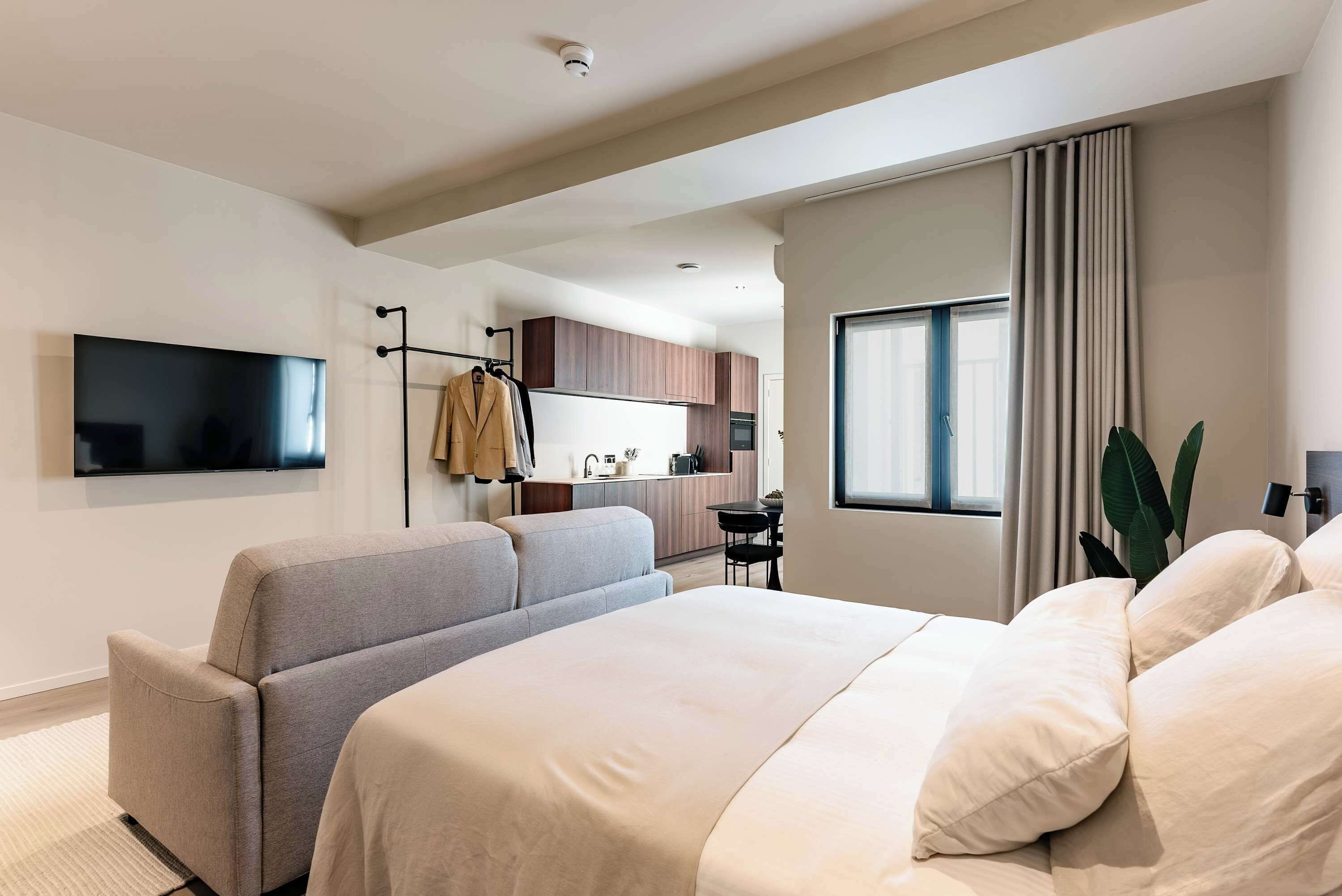 A modern hotel room with a large bed, a seating area, a flat-screen TV, a kitchenette, and a window with curtains.