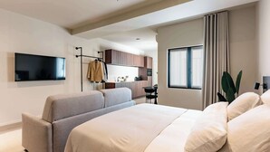 Habitación de hotel moderna con una cama grande, zona de estar, televisor de pantalla plana, kitchenette y ventana con cortinas.