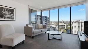 A modern living room with a sofa, armchair, and coffee table, large windows with a city view, and a framed picture on the wall.