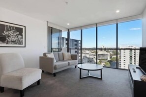A modern living room with a sofa, armchair, and coffee table, large windows with a city view, and a framed picture on the wall. - ML1306A Milton1306 ; 1 Bedroom 1 Bathroom (Brisbane)