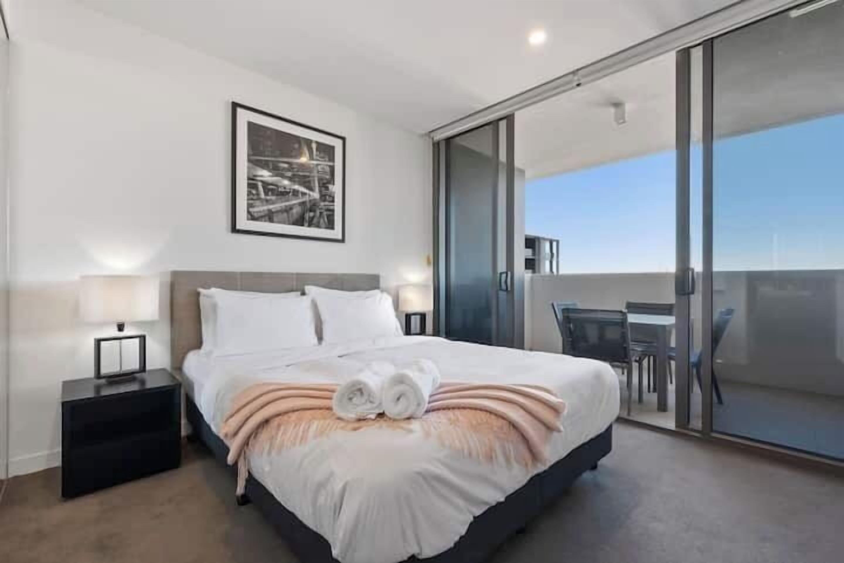 Une chambre d’hôtel moderne avec un grand lit, une table de chevet, un tableau encadré au mur et une porte-fenêtre coulissante donnant sur un balcon équipé de mobilier d’extérieur.