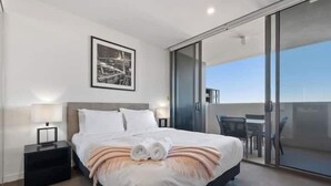 Ett modernt hotellrum med en stor säng, ett nattduksbord, en inramad tavla på väggen och en skjutdörr i glas som leder ut till en balkong med utomhusmöbler.