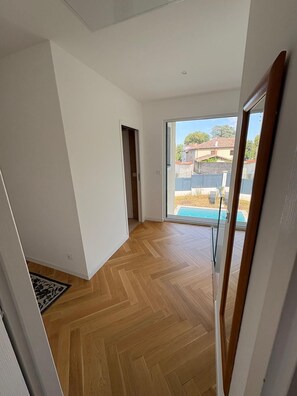 A room with a wooden herringbone floor, a sliding glass door leading to a pool, and a view of neighboring houses. - House between the city and the countryside near Lyon (Saint-Genis-Laval)