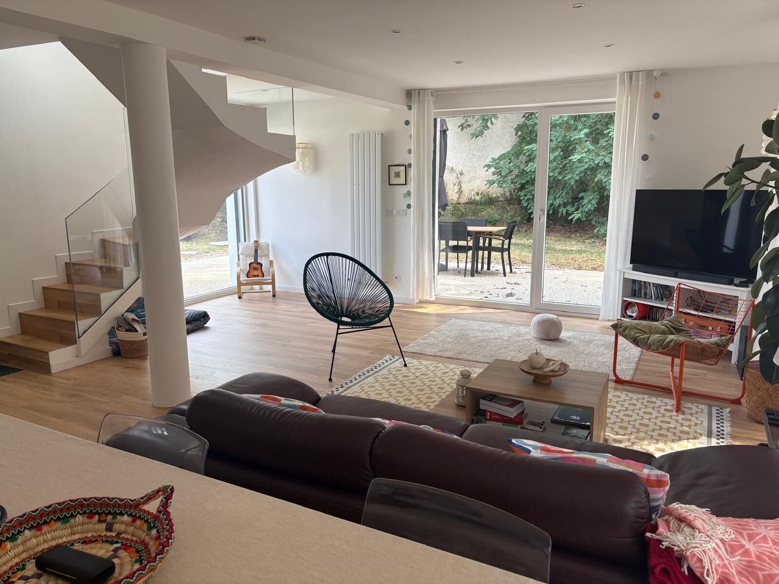 A modern living room with a staircase, a black chair, a sofa, a television, and a large window.