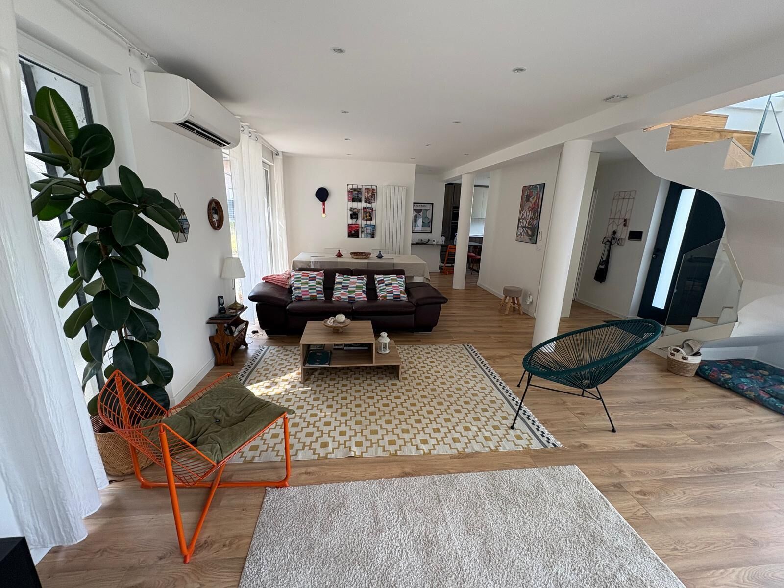 Un salon moderne avec un canapé, une table basse et des fauteuils, une grande plante et un escalier menant à un niveau supérieur.