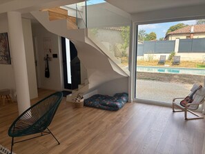 Un interior modern amb una escala que condueix a un balcó, una zona d'estar amb una cadira i una banca, i vista a una piscina a l'exterior.