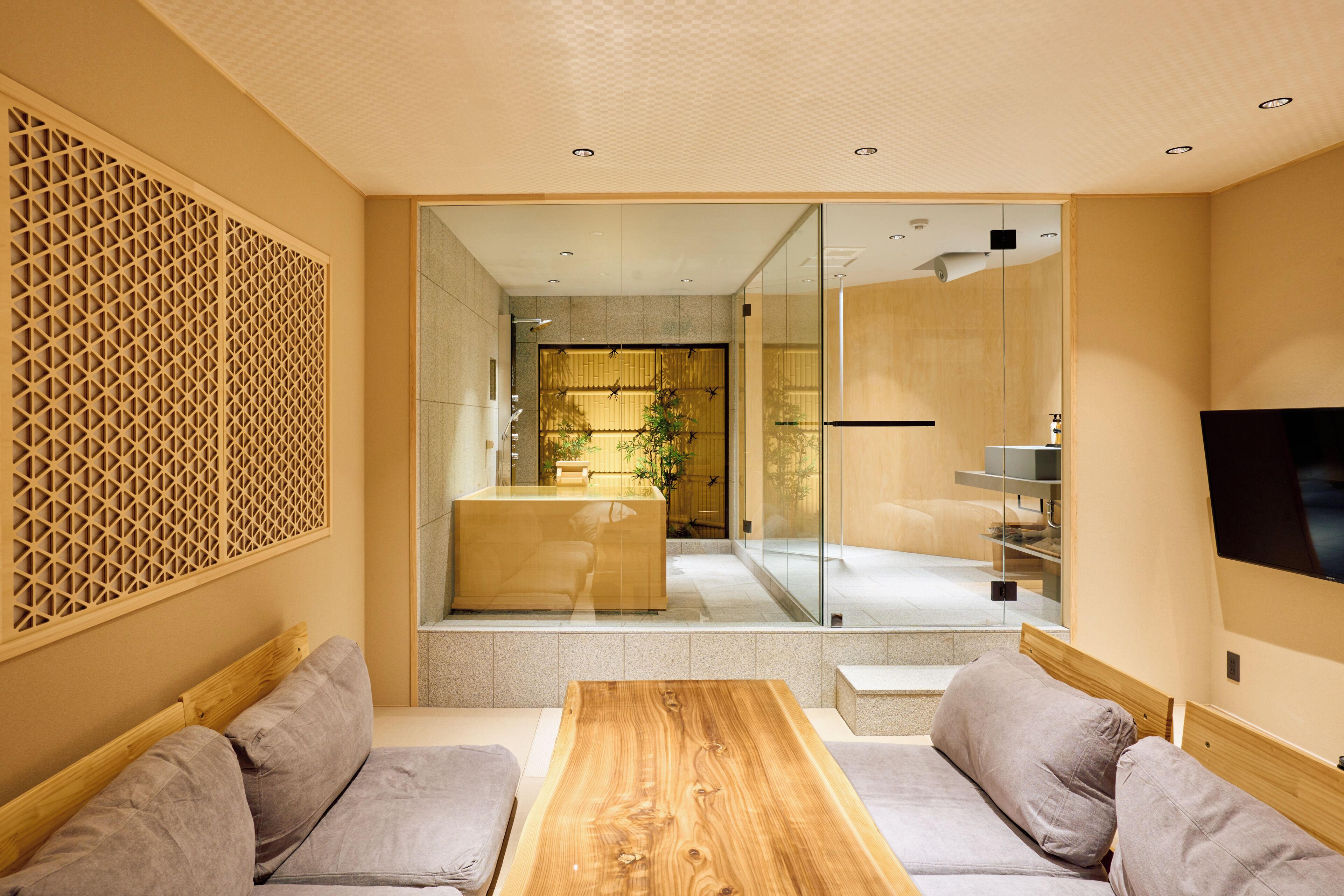 A modern living room with a wooden coffee table, grey sofas, and a glass partition leading to a kitchen area.