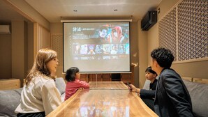 A group of people seated around a wooden table in a room with a projection screen displaying a Netflix interface. - Imakusa-so, Osaka: Tempozan - Ikisu - (大阪府大阪市港区築港3丁目10−11)