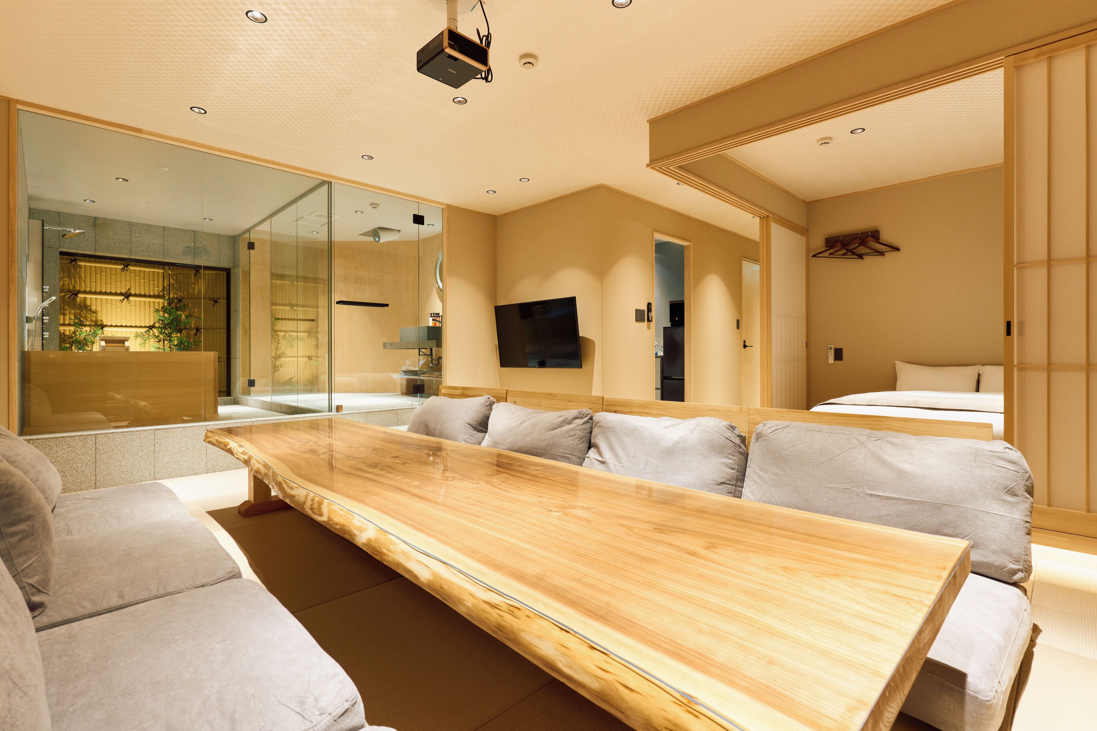 A modern hotel room with a long wooden table, a sofa, a TV, and a large mirror.
