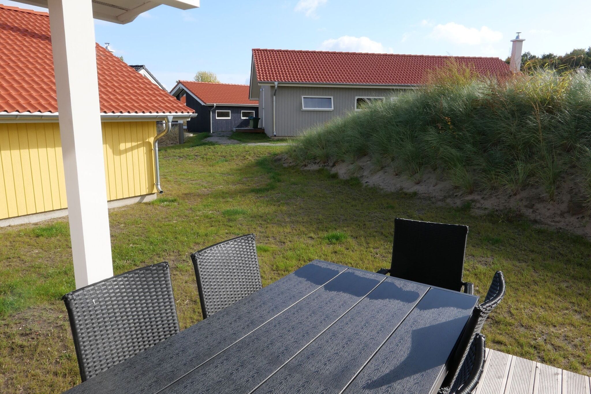 An outdoor patio with a table and chairs, surrounded by a grassy area and a building with a red roof.