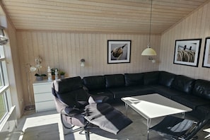 Un salon moderne avec un canapé d’angle en cuir noir, une table basse blanche et une petite table d’appoint. Deux lampes murales et des tableaux encadrés ornent le mur.