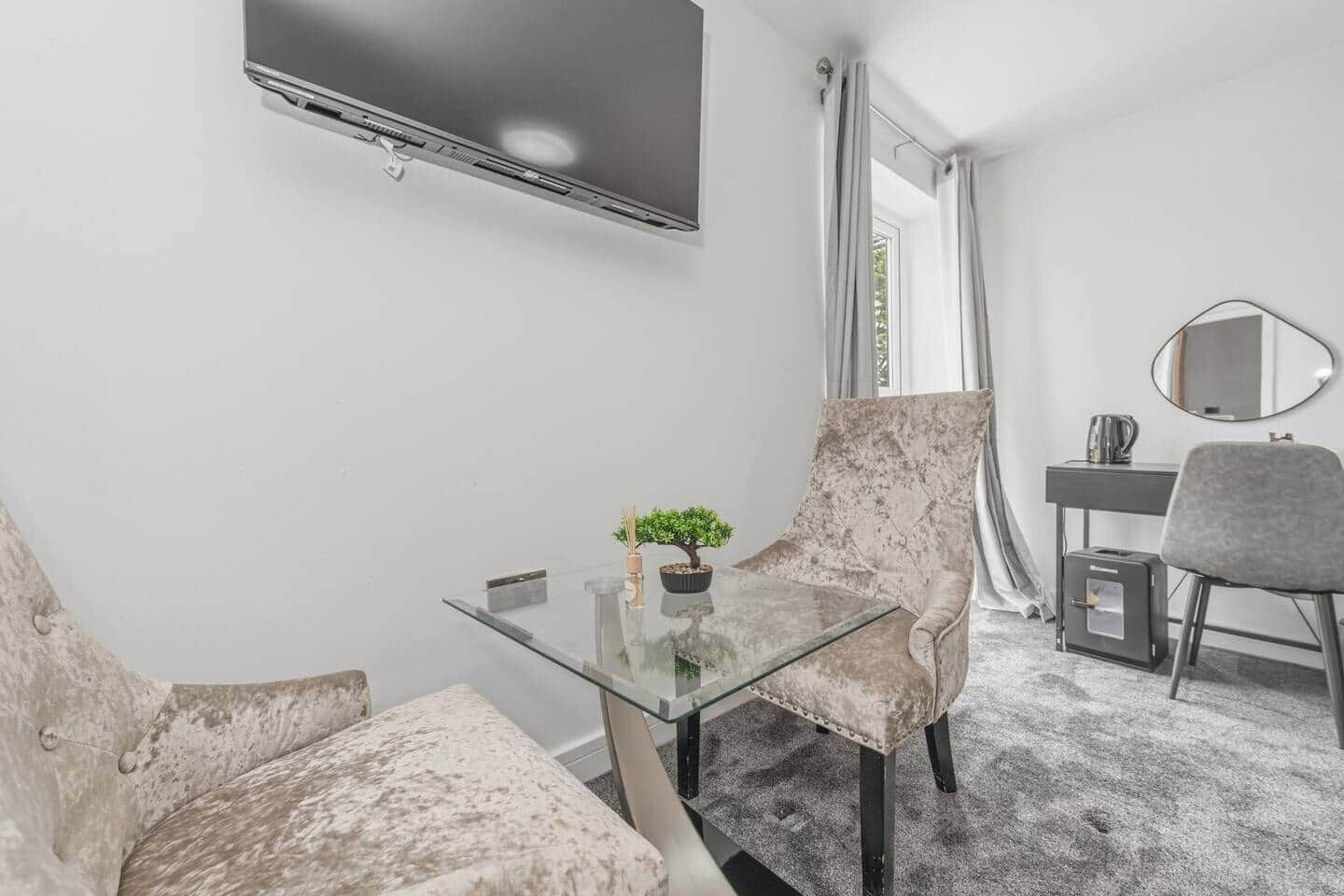 Een moderne kamer met een glazen tafel, twee comfortabele stoelen, een kleine potplant, een flatscreen televisie aan de wand, een bureau met stoel, en een spiegel.