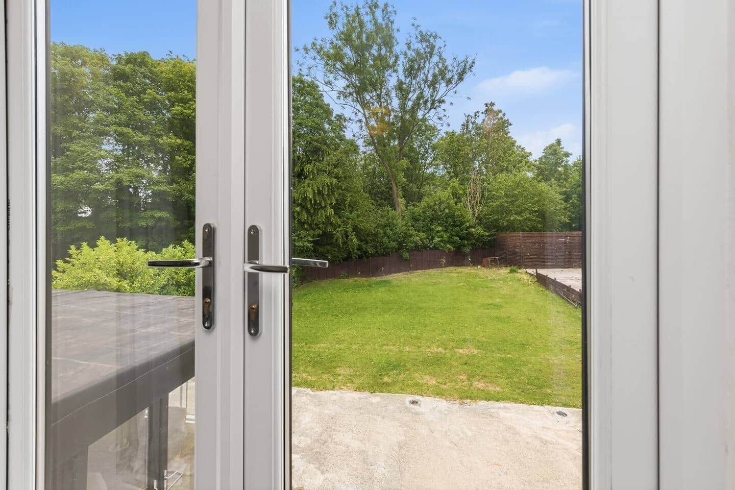 A view through a glass door into a backyard with a grassy lawn, trees, and a brick wall.