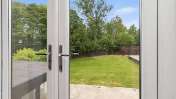A view through a glass door into a backyard with a grassy lawn, trees, and a brick wall.