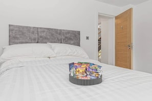 Un lit bien fait, avec du linge de lit blanc et une tête de lit grise, une table de chevet sur laquelle sont disposés divers en-cas, et une porte en bois à l’arrière-plan.