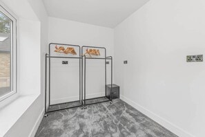 Une pièce d’angle avec une fenêtre, un tapis gris, un étendoir métallique sur lequel sont suspendus des vêtements et une petite armoire.