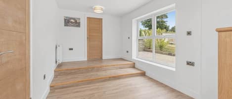 Un pasillo con piso de madera, una puerta de madera, una ventana con vista a follaje y una lámpara en el techo.