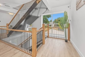 Una escalera con barandilla de madera y balaústres metálicos, que conduce a un espacio exterior con vistas a zonas verdes y un cielo despejado.