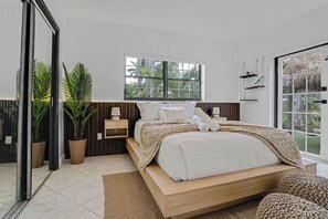 Ein modernes Schlafzimmer mit einem großen Bett, einem Nachttisch, einem Beistelltisch und einem Fenster mit Blick auf Grünflächen.