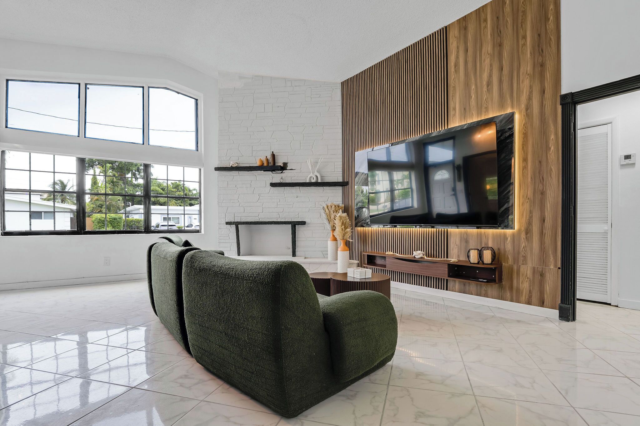 Uma sala de estar moderna com uma grande televisão de tela plana, um sofá estofado verde e uma parede de tijolos brancos.