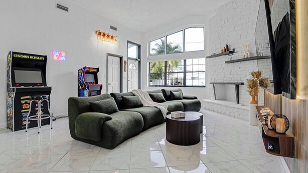 A modern living room with a green sofa, arcade games, and a large window. - Waterfront House with Tiki & Firepit (Miami)