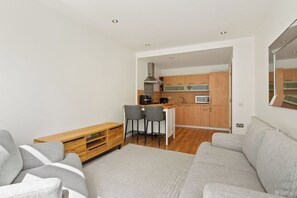 A modern living room with a grey sofa, a wooden coffee table, and two grey chairs. A kitchen with wooden cabinets and a microwave is visible through an open doorway. - Modern City Centre Apt with Balcony in Dublin 1 (Dublin)