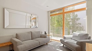 Ein modernes Wohnzimmer mit einer Couch, einem Sessel, einem großen Spiegel und einem Fenster mit Blick auf Bäume.