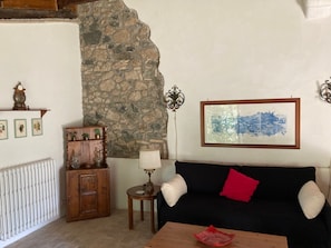A living room with a stone wall, a wooden cabinet, a black sofa with a red pillow, a framed picture, and a small table with a red object on it. - The Casalino Orvieto Dépendance. A hidden oasis surrounded by greenery, 15 minutes from Orvieto (Umbria)