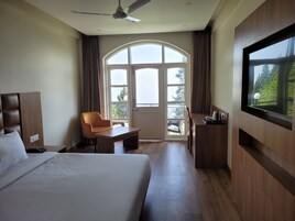 Habitación de hotel con cama, escritorio, silla, televisor y una ventana grande con vistas a la vegetación.