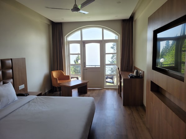 A hotel room with a bed, a desk, a chair, a television, and a large window with a view of greenery.