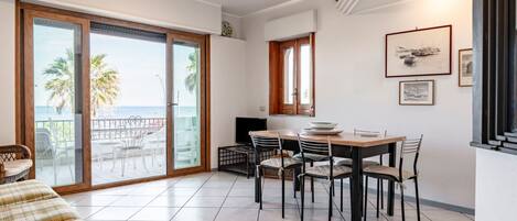 A dining area with a table and chairs, a sofa, and a view of the sea through a sliding glass door.