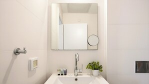 Et badeværelse med en hvid vask, et spejl, en vandhane, en potteplante og toiletartikler på bordet.