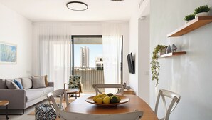 En moderne stue med et spisebord, stole, en sofa, et fjernsyn og en balkon med udsigt til bygninger.