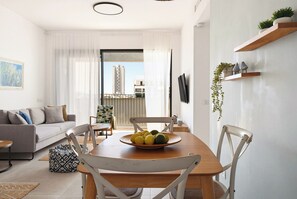 A modern living room with a dining table, chairs, a sofa, a TV, and a balcony with a view of buildings. - Bright Spacious Stay with Balcony (Tel Aviv)