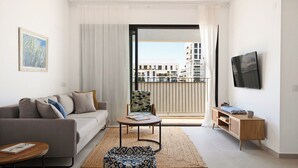 En moderne stue med sofa, et lille bord, en mønstret puf, et fjernsyn og en balkon med udsigt til bygninger.