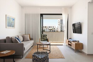 A modern living room with a sofa, a small table, a patterned ottoman, a TV, and a balcony with a view of buildings. - Bright Spacious Stay with Balcony (Tel Aviv)