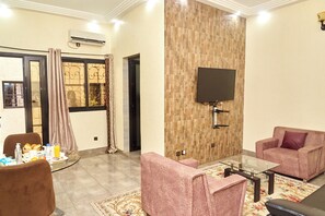 Una sala de estar con una televisión de pantalla plana montada en una pared de madera, dos sillones rosa, una mesa de centro de vidrio y un comedor con una bandeja de desayuno.