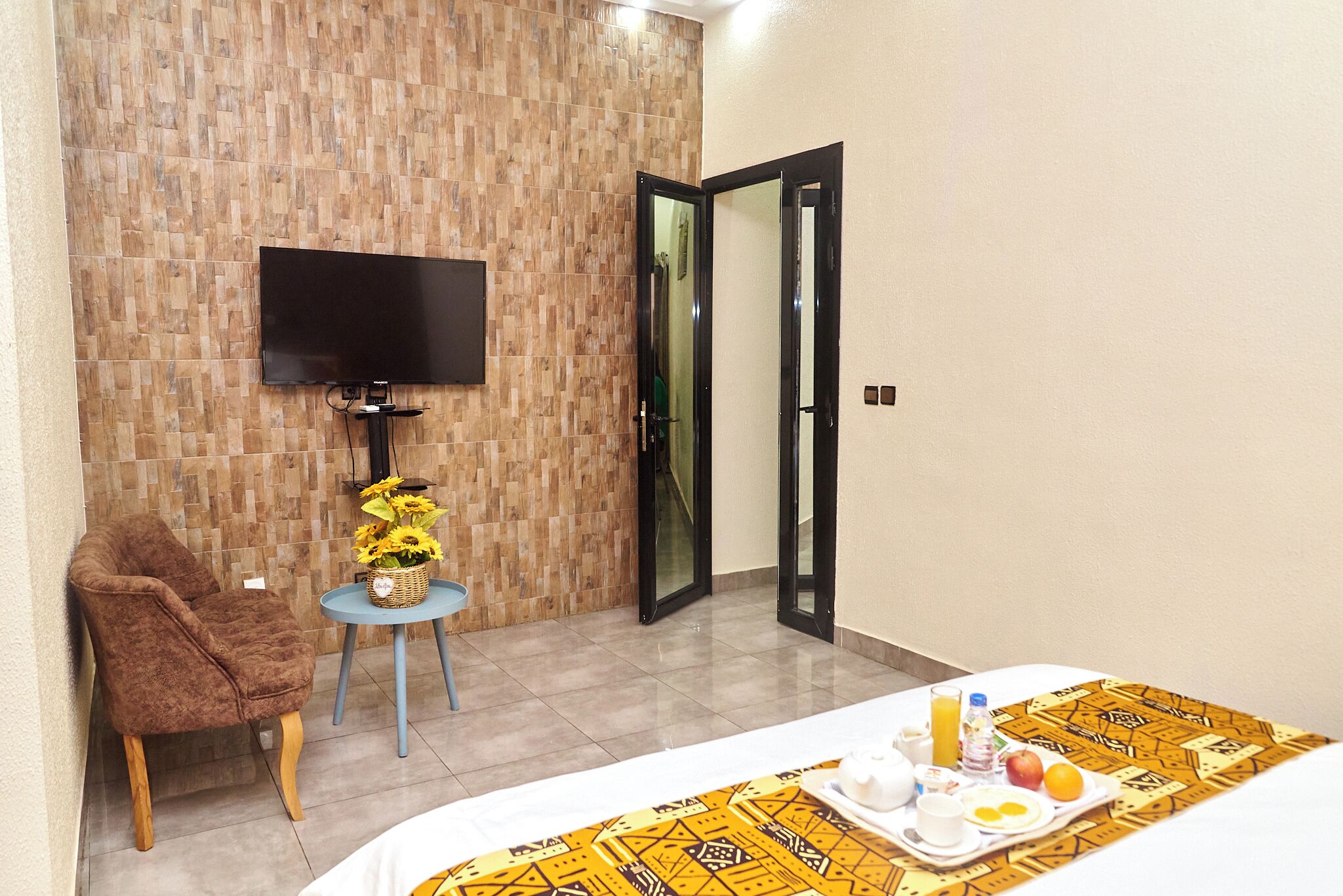 Quarto de hotel com cama, televisão na parede, mesa pequena com um vaso de flores e uma cadeira.