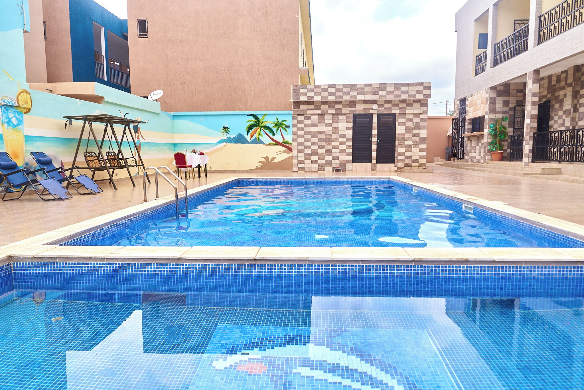 Piscina com parede xadrez, cadeiras de espreguiçadeira e mesa com mural de praia.