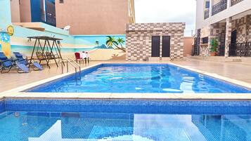 Piscina com parede xadrez, cadeiras de espreguiçadeira e mesa com mural de praia.