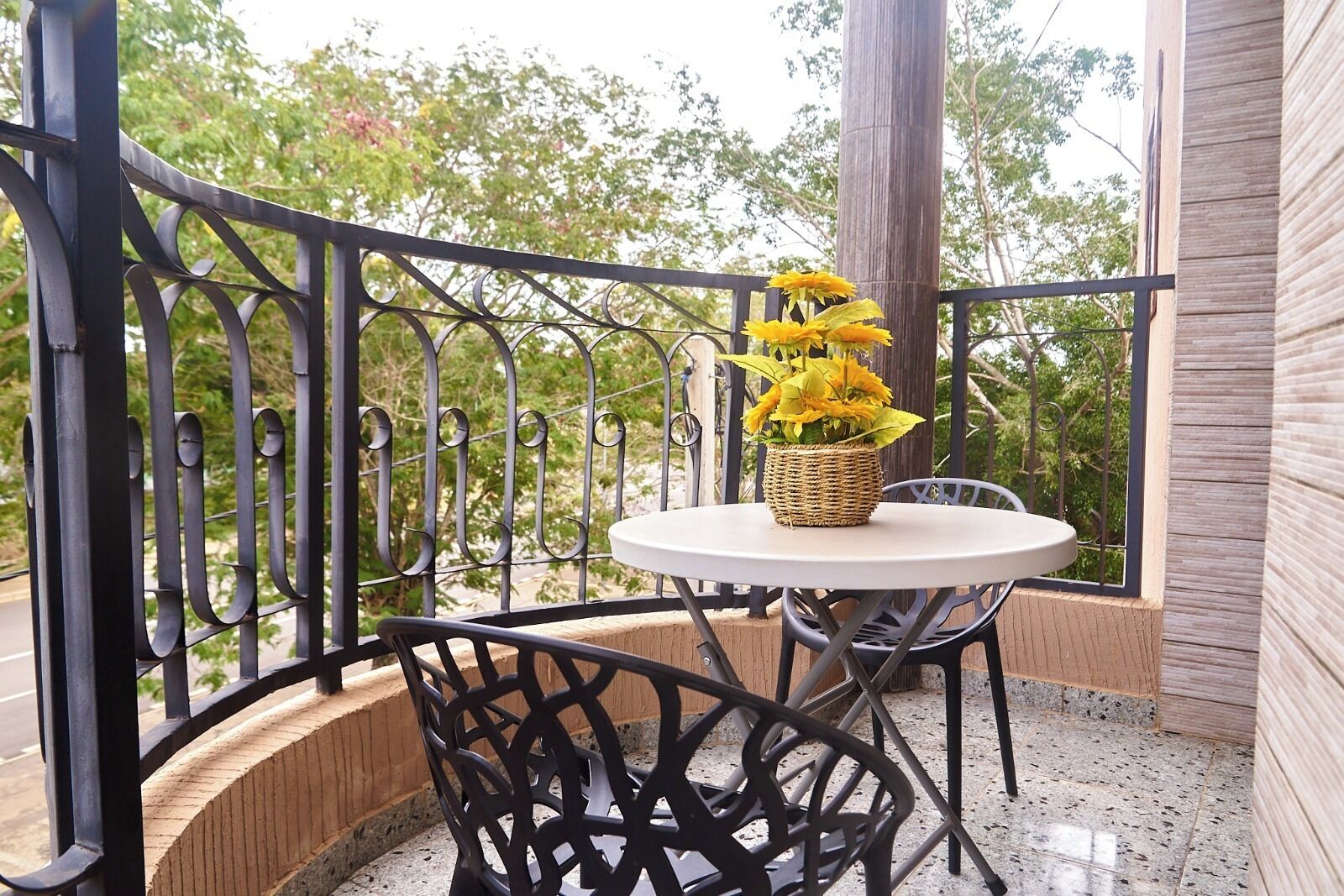 Uma varanda com uma mesa redonda, um cesto de vime com flores amarelas e cadeiras de metal.