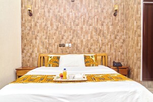 Quarto de hotel com parede em padrão de madeira, cama com colcha branca e almofadas estampadas, mesa de cabeceira com telefone e bandeja com itens de café da manhã.
