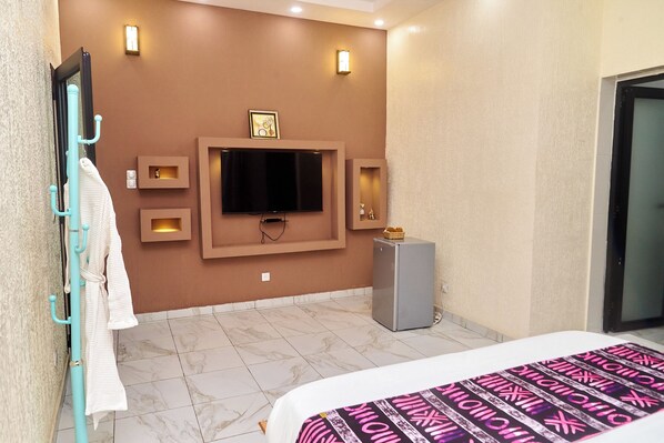 A hotel room with a flat-screen TV mounted on the wall, a small cabinet, a bed with a patterned bedspread, and a bathroom door.
