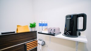 Habitación de hotel con escritorio de madera, cafetera negra, estante con botellas de agua y tazas, control remoto y toma de corriente.