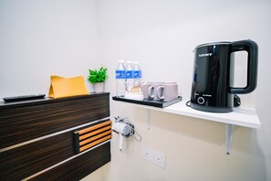 Habitació d'hotel amb escriptori de fusta, cafetera negra, prestatges amb ampolles d'aigua i gots, comandament a distància i presa elèctrica.