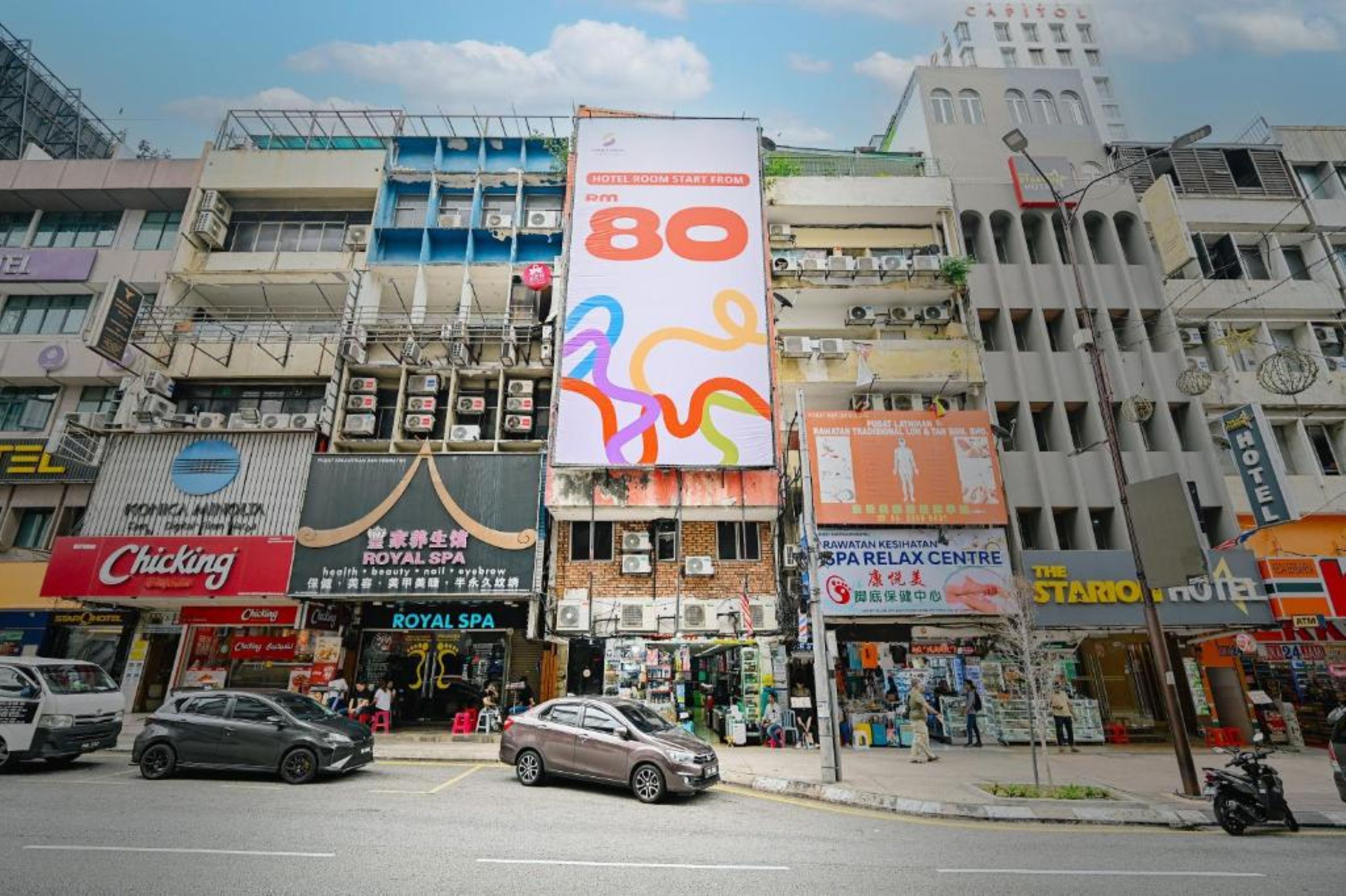 Une vue de rue avec plusieurs immeubles commerciaux, dont un hôtel dont l’enseigne bien visible annonce des prix à partir de 80 RM.